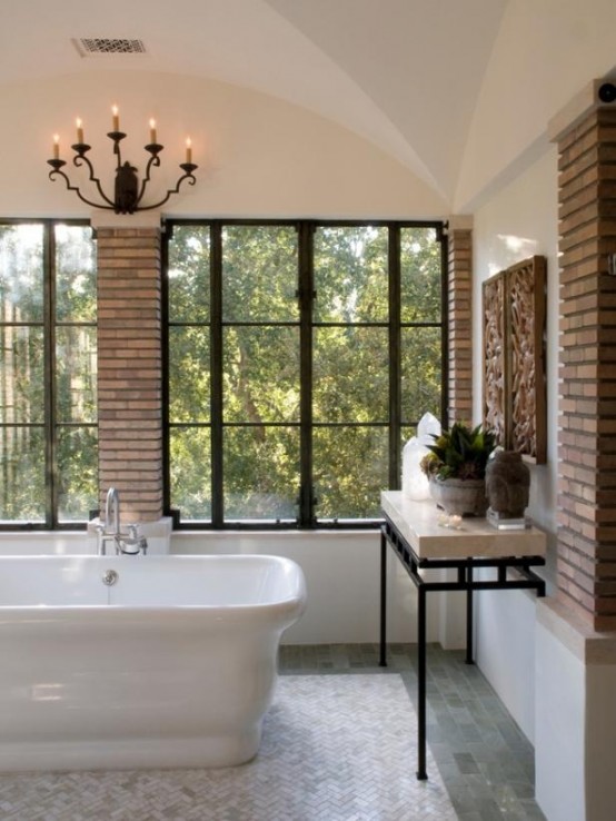 a modern bathroom with brick walls, large framed windows, a tiled floor, a large tub and a chic chandelier