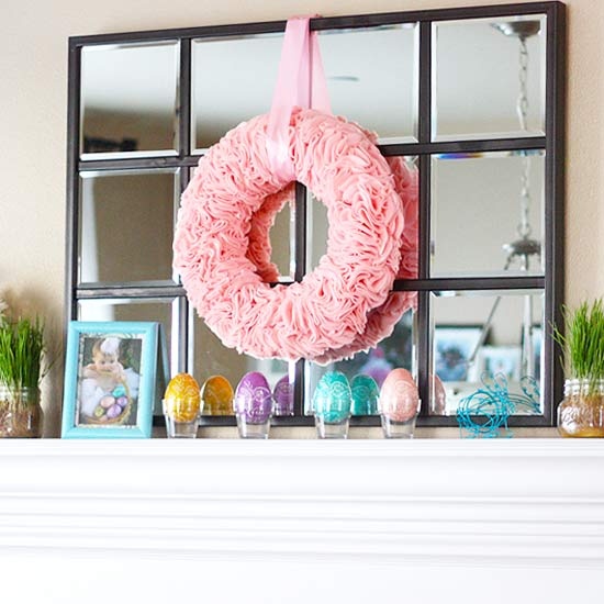 a colorful easter mantel with a pink wreath, colorful eggs on stands and some potted grass