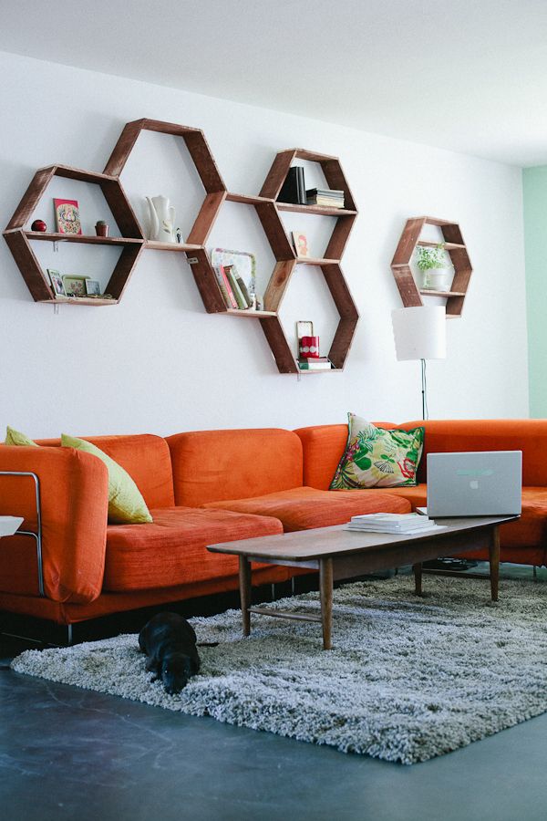 a living room with an orange sectional sofa, a stained hexagon shelf on the wall, a grey rug and a stained coffee table plus a printed pillow is cool