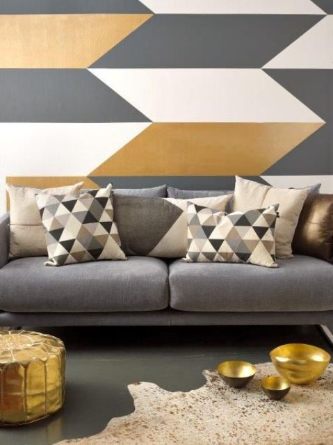 an elegant modern living room with a chevron printed accent wall, a grey sofa with geo printed pillows, a gold pouf and a neutral rug with gold bowls