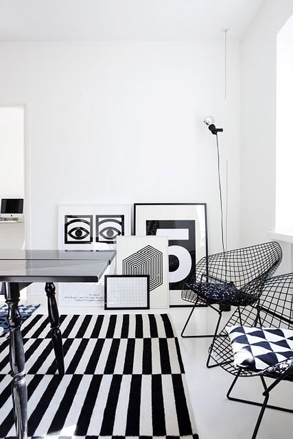 a black and white living room with a striped rug, a vintage black table, black wire chairs with black and white geo print cushions, a gallery wall right on the floor