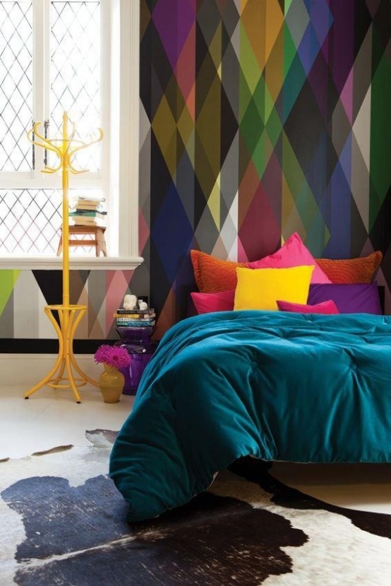 a colorful bedroom with a super bold geometric wall, a bed with colorful pillows and a blanket, a purple side table and a yellow rack
