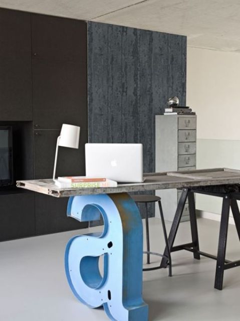 a home office with a metal clad fireplace, a rough wood wall, an industrial desk of metal and reclaimed wood and a blue metal monogram that holds the desktop