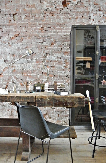 an eclectic home office with an exposed brick accent wall, a wood and glass storage unit, a rough wood desk that feels shabby and industrial and leather chairs
