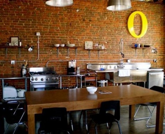 a fully industrial kitchen with a red brick wall and shiny metal surfaces plus a bold yellow letter and a wooden table