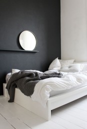 a minimalist bedroom with a black accent wall, a white bed with monochromatic bedding, a shelf with a round mirror and nothing else