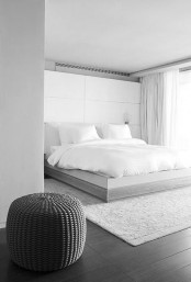 a stylish minimalist bedroom in grey and white, with a sleek storage unit behind the bed, a sleek bed with white bedding and a neutral rug and a pouf