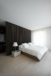 a minimalist bedroom with a walk-in closet hidden with a large dark curtain and a neutral bed with white bedding, a sleek nightstand with a white lamp and much natural light