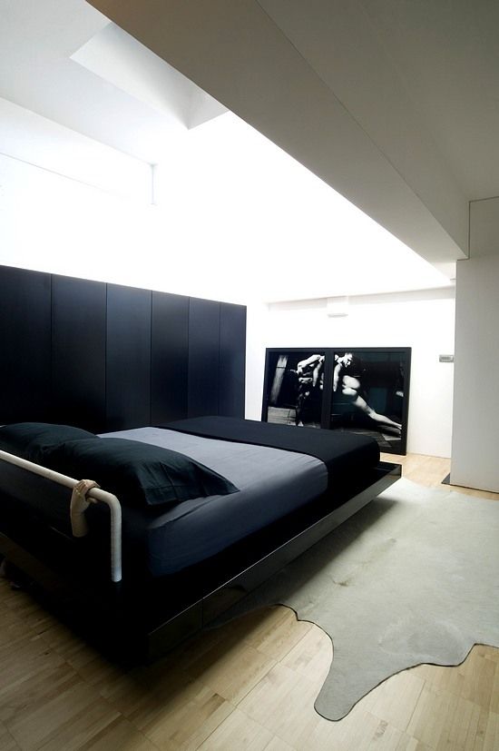a minimalist bedroom with a sleek black storage unit, a glossy black bed with grey and black bedding, a statement artwork and a grey rug plus lots of natural light