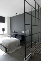 a Scandi meets minimalist bedroom with a grey accent wall, a grey bed with a storage headboard, black bedding and black lamps plus a glass space divider