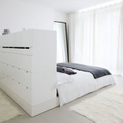 a minimalist white bedroom with a bed with a storage headboard, with lots of drawers, a floor mirror and a glazed wall with curtains