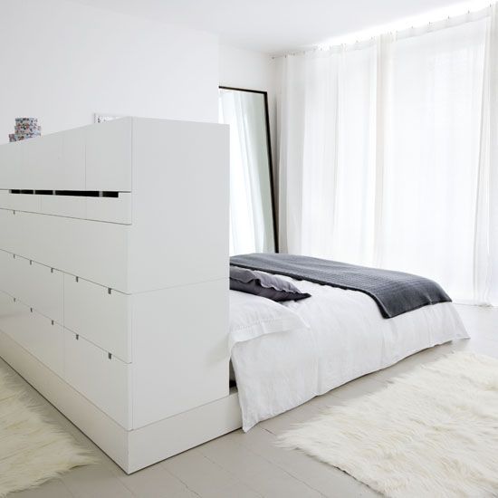 a minimalist white bedroom with a bed with a storage headboard, with lots of drawers, a floor mirror and a glazed wall with curtains