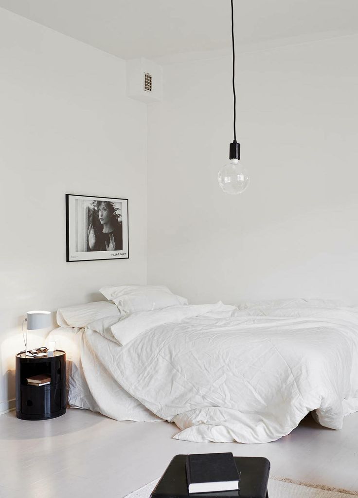 a Scandinavian meets minimalist bedroom with a bed with white bedding, a sleke black nightstand, some bulbs, a black stool and an artwork