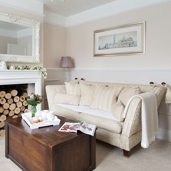 a beautiful and refined neutral living room with a fireplace with firewood stacked, a neutral patterned sofa, a dark chest as a coffee table, an artwork and a mirror in an ornated frame