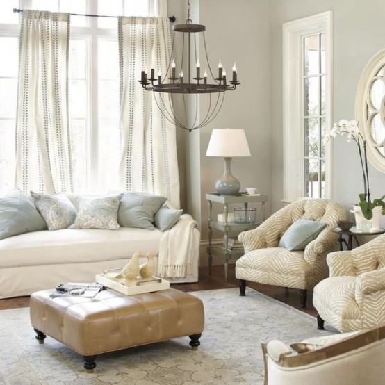 a chic neutral living room with grey walls, neutral seating furniture, a leather upholstered ottoman, a vintage chandelier and flowy curtains is very stylish