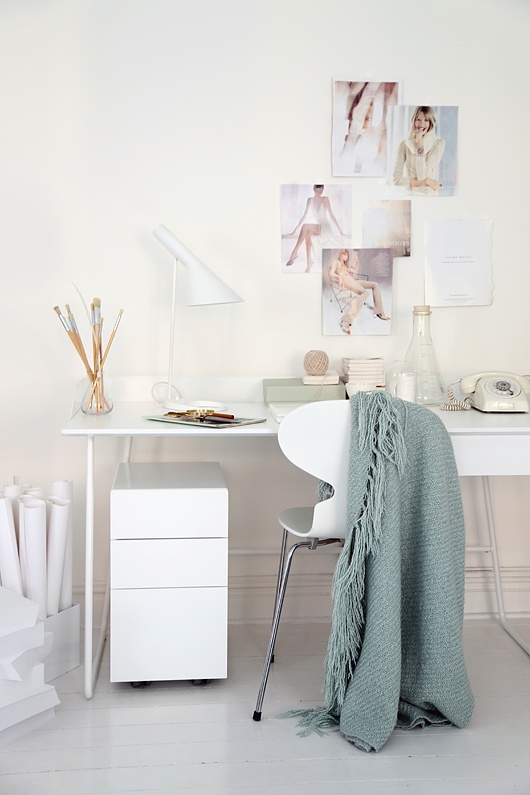 a white Scandinavian home office wiht a desk, chair, a cabinet, a table lamp, a gallery wall and a green blanket is a cool idea