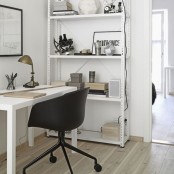 a small white Scandinavian home office with a large open shelving unit, a desk, a black chair, a brass table lamp and some artwork