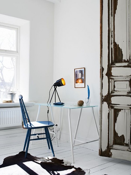 an airy white Scandinavian home office with a glass and metal desk, a blue chair, some lovely decor and a black table lamp