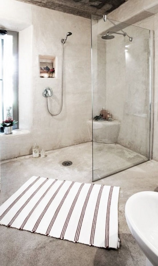 a fully concrete bathroom with a shower space divided with a glass partition, a striped rug and a rough wood ceiling