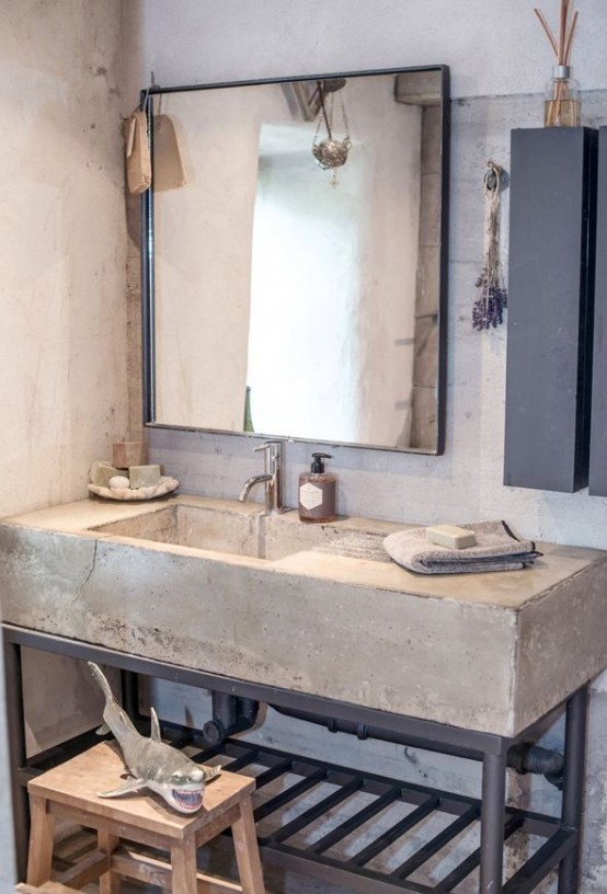 an industrial bathroom with concrete walls and a cocnrete sink, a wooden stool and a framed mirror is a relaxed space