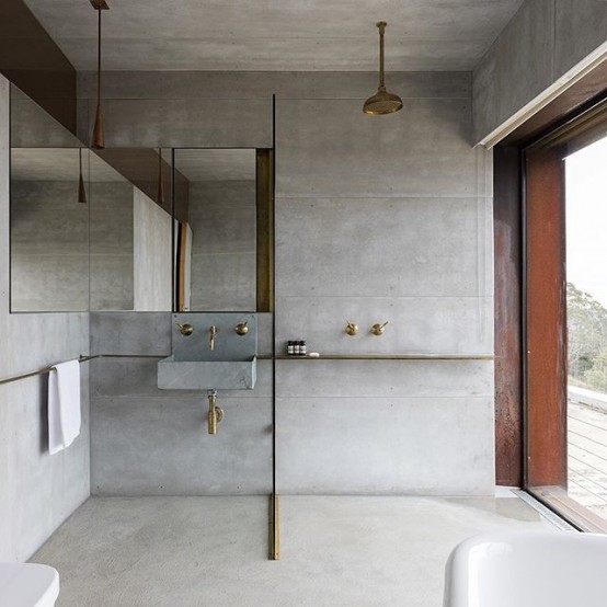 a minimalist concrete bathroom with a large mirror, brass fixtures and a concrete sink plus a glazed wall with a view
