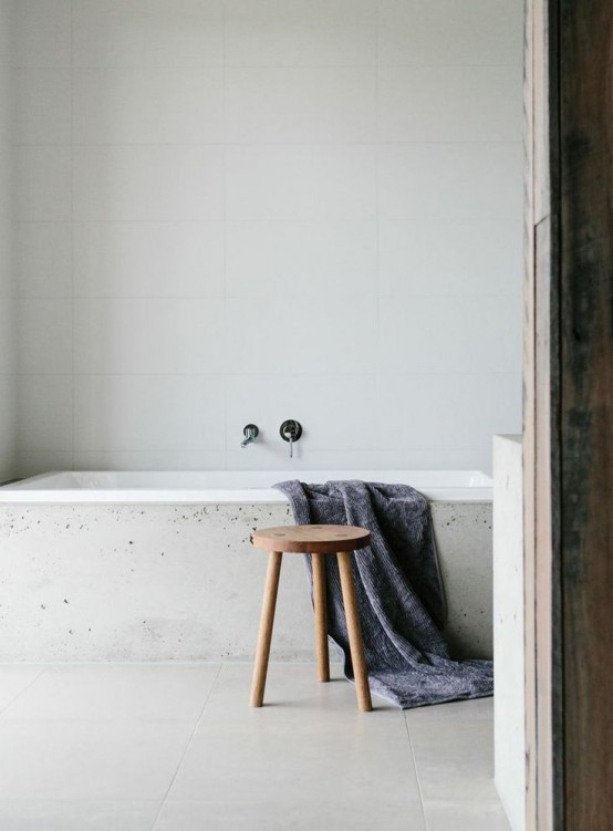 a stylish and minimalist bathroom with white tiles all over the space and a bathtub clad with concrete is airy and chic