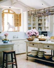 a whimsy cottage kitchen with white cabinets and a shabby chic kitchen island, with a blue backsplash, wooden shutters and a creative hanger over the island