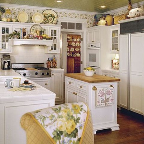a lovely cottage kitchen with a yellow ceiling, white cabinets and a compact kitchen island, a floral wallpaper accent, decorative porcelain and floral textiles
