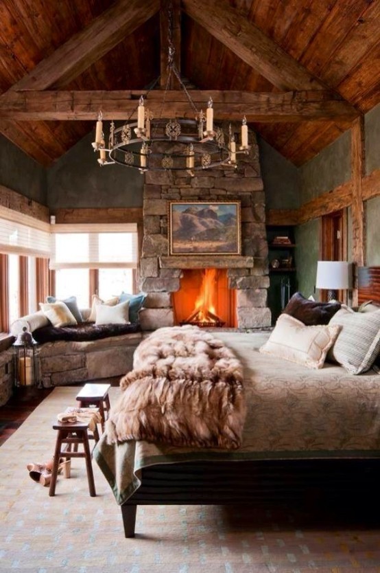 a cozy chalet bedroom with a wood clad ceiling, a stone fireplace and a corner bench of stone, a bed with cozy bedding and a metal chandelier