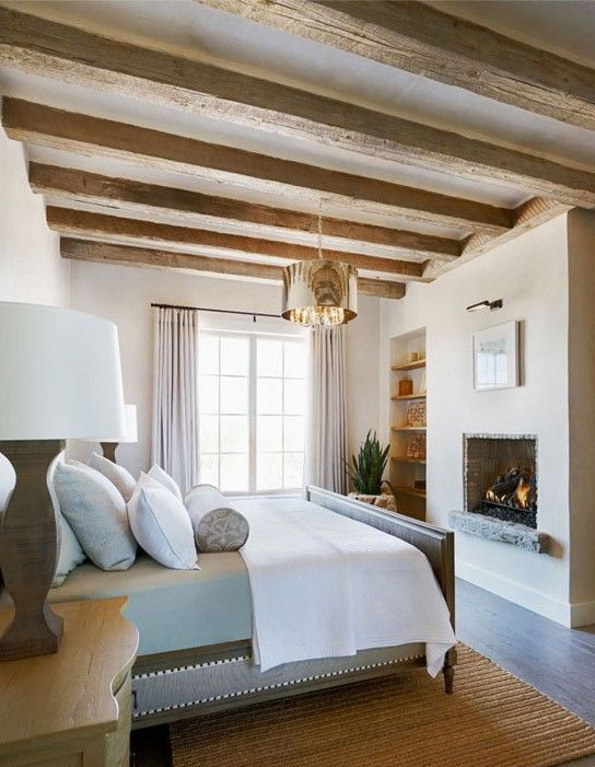 a stylish and chic bedroom with wooden beams, a bed with neutral bedding, a fireplace and built-in shelves plus lamps