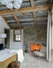 a modern chalet bedroom with stone walls and a slanted ceiling, a bed with neutral bedding, a neutral chair and a clear glass nightstand