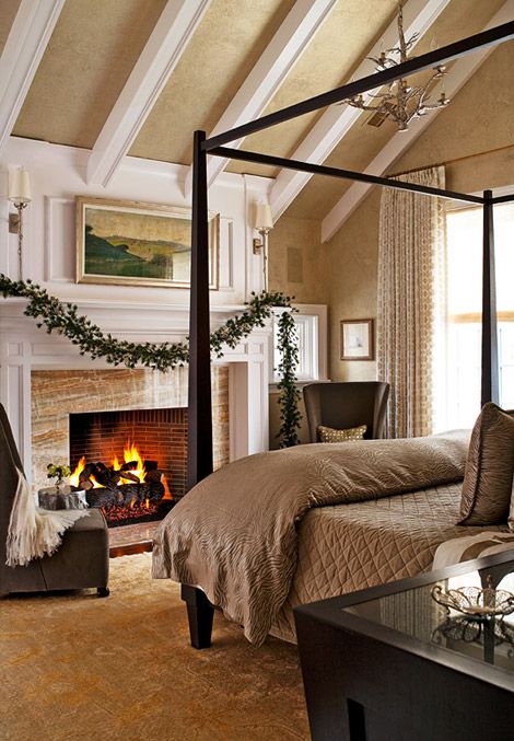an elegant tan bedroom with a fireplace, a frame bed with beige bedding, taupe chairs and some greenery