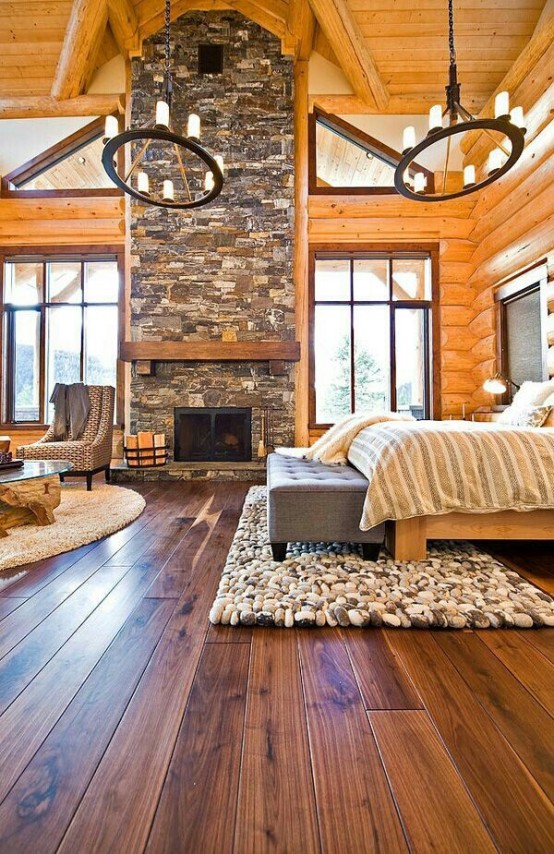 a welcoming chalet bedroom with wood clad walls, a stone fireplace, a bed with neutral bedding, a pouf and statement chandeliers