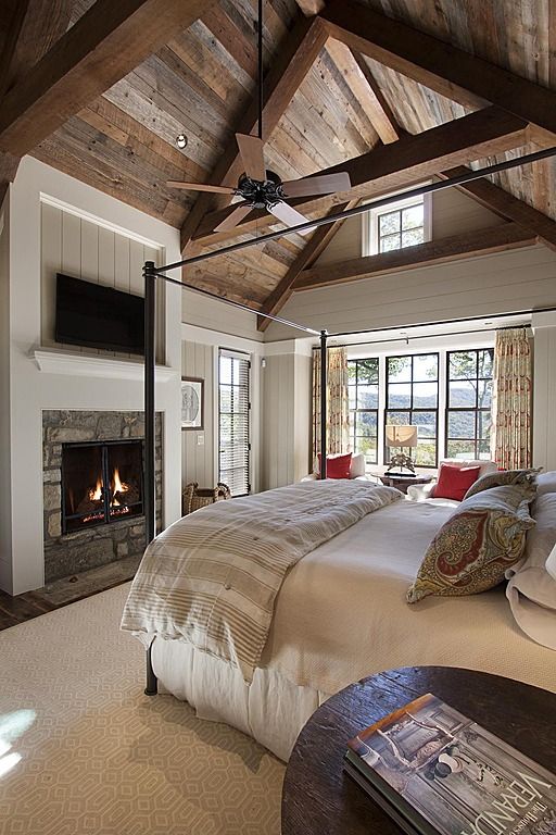 a chalet bedroom with an attic ceiling done with wood, a stone fireplace, a frame bed, printed bedding and a sitting zone by the window