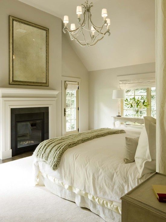 a stylish neutral bedroom with a bed and neutral bedding, a fireplace, a chandelier and a large mirror over the fireplace