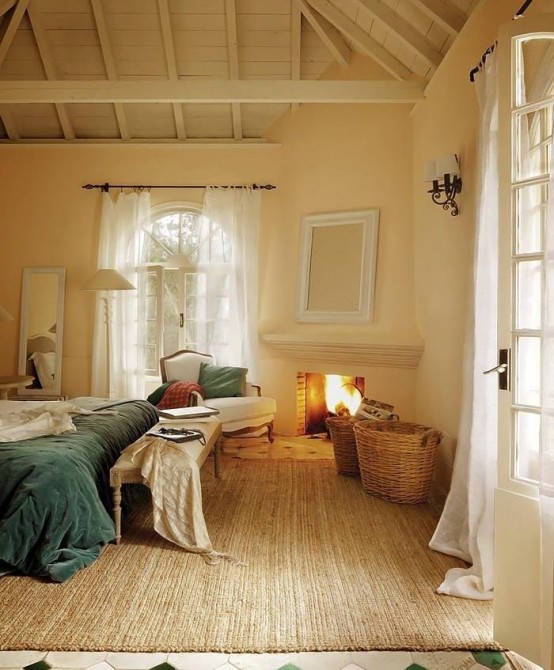 a French cottage bedroom with a fireplace, a bed with blue bedding, a bench with blankets and a basket with various stuff stored