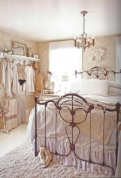 a neutral and refined shabby chic bedroom with printed wallpaper, a forged bed, an open wardrobe and a vintage chandelier and exquisite accessories
