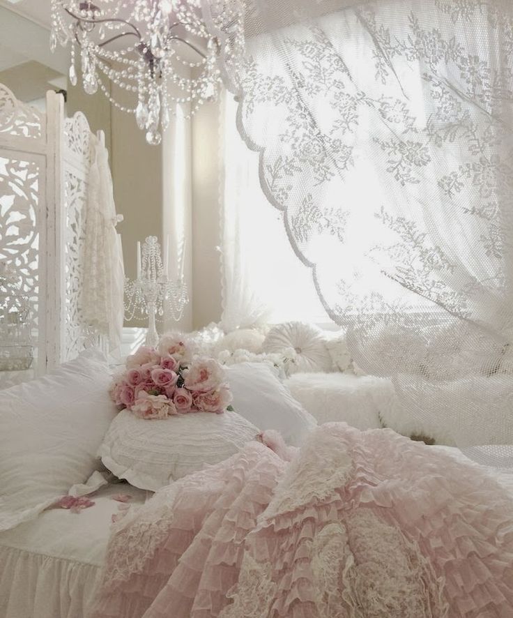 a white shabby chic bedroom with exquisite white furniture, a carved screen, a crystal chandelier, blush bedding and a lace curtain