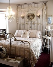 a shabby chic bedroom with a forged bed, a wooden headboard, artworks, a crystal chandelier and neutral bedding