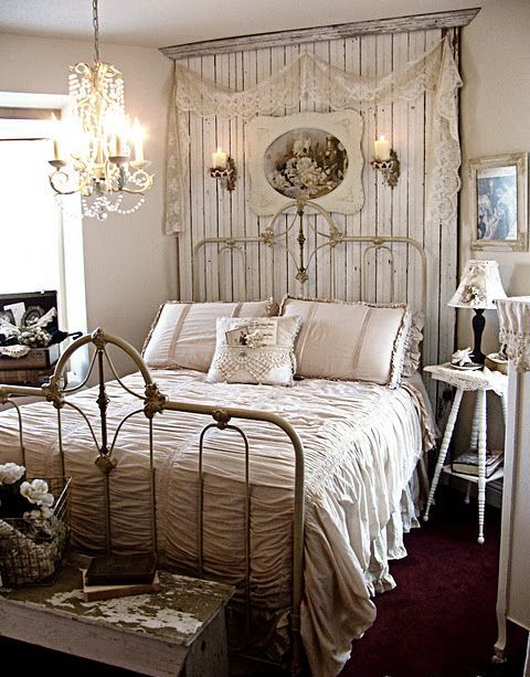 a shabby chic bedroom with a forged bed, a wooden headboard, artworks, a crystal chandelier and neutral bedding
