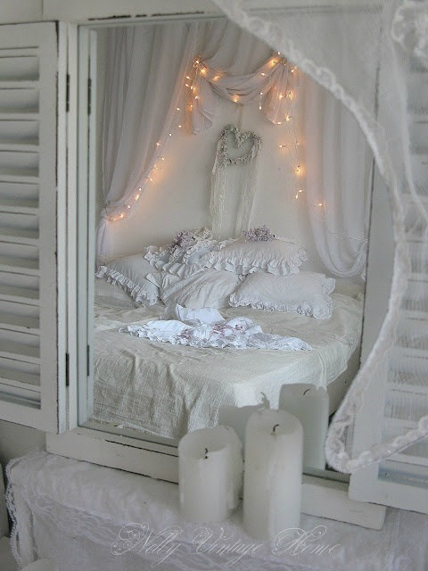 a white shabby chic bedroom with shutters and a mirror, a bed with refined ruffle and floral bedding and lights over the bed