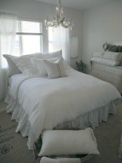 a white shabby chic farmhouse bedroom with refined furniture, a crystal chandelier, white bedding and potted greenery