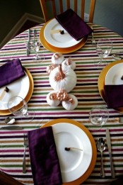 a bright Thanksgiving tablescape with a striped tablecloth, gold chargers, deep purple napkins and white pumpkins with copper stems