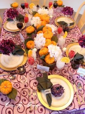 a colorful Thanksgiving table setting with a purple printed tablecloth, purple and white blooms, natural pumpkins and leaves, a purple pumpkin