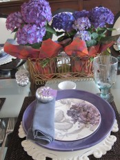 a vintage-inspired purple Thanksgiving table setting with purple blooms and plates, brown placemats, silver pumpkins and refined white chargers
