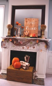 a rustic Thanksgiving mantel with vines, faux leaves, wheat bundles, candles, pumpkins on stands and a wooden sign plus some firewood and pumpkins