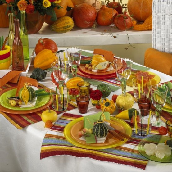 a colorful Thanksgiving tablescape with bold striped placemats, colored glasses, bottles and candleholders, some gourds and pumpkins is amazing