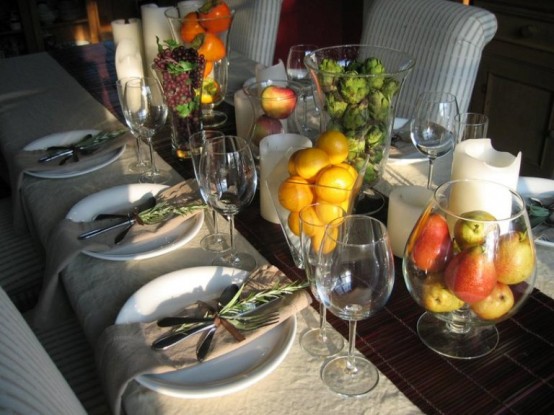 a chic fall or Thanksgiving tablescape with neutral linens, a brown table runner, bold fruit in tall glasses, large pillar candles and greenery that accents each place setting