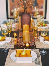 a chic warm-colored Thanksgiving table setting with amber glasses and plates, with pillar candles, white porcelain and woven placemats plus faux branches
