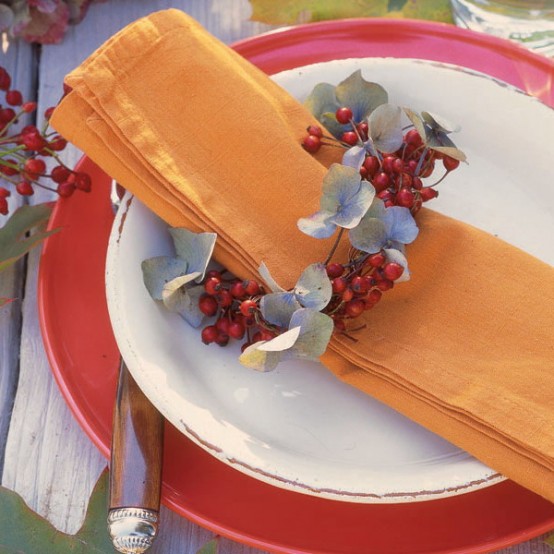 a bold place setting with a red charger, an orange napkin with a ring of berries and pale greenery for a chic and bold fall-inspired look